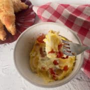 Bowl of baked eggs with tomatoes and herbs next to a plate of croissants and a red checkered napkin; a fork holds a bite of the egg mixture.