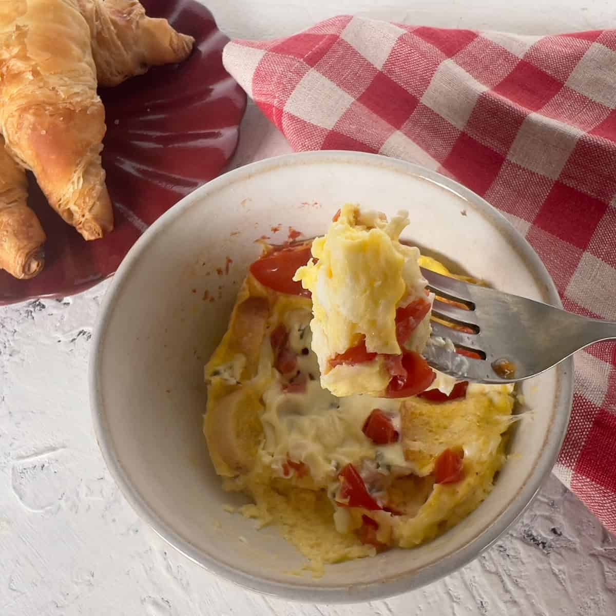 Bowl of microwaved eggs with tomatoes next to a plate of croissants and a red checkered napkin; a fork holds a bite of the quiche.