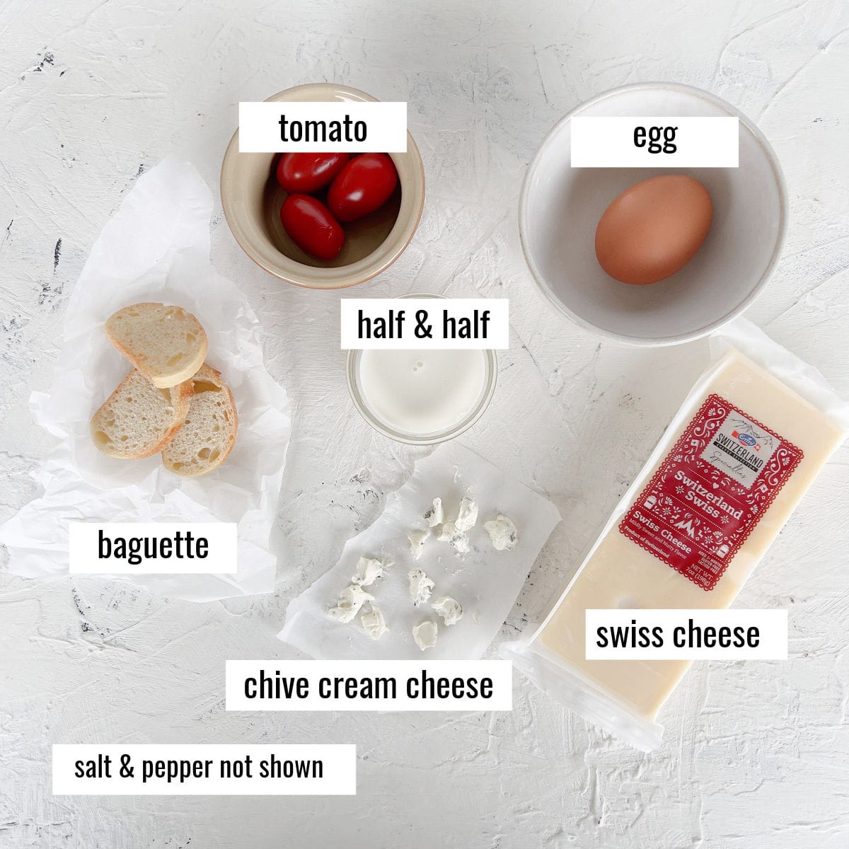 Ingredients for a quiche in microwave are arranged on a white surface: tomato, egg, half & half, Swiss cheese, chive cream cheese, and baguette slices.