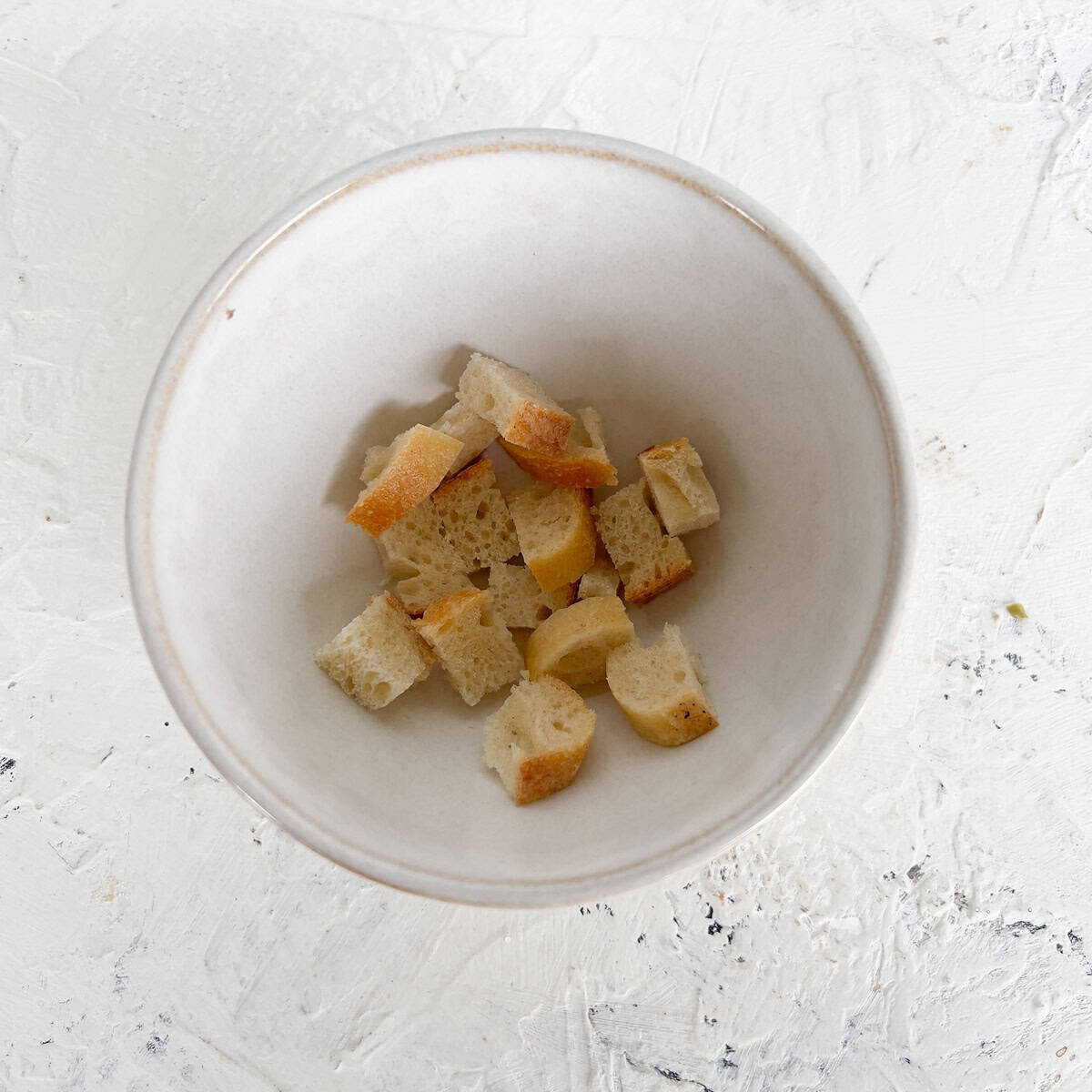 A white bowl containing several small cubes of bread, sits on a light textured surface.