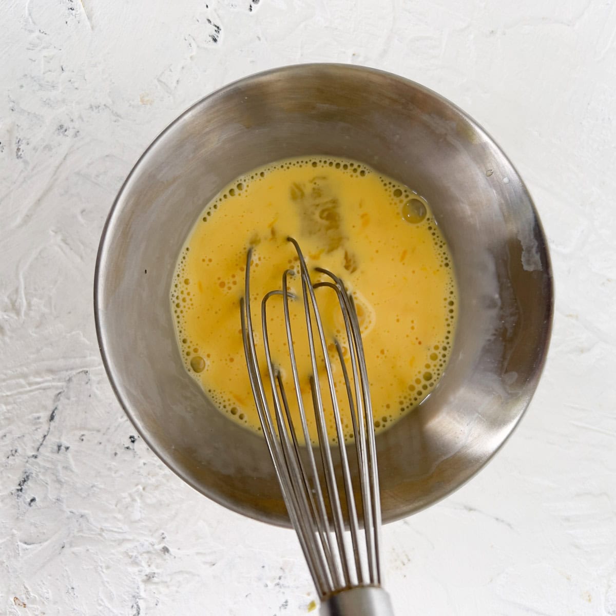 A metal whisk is resting in a metal bowl containing beaten eggs on a white textured surface.