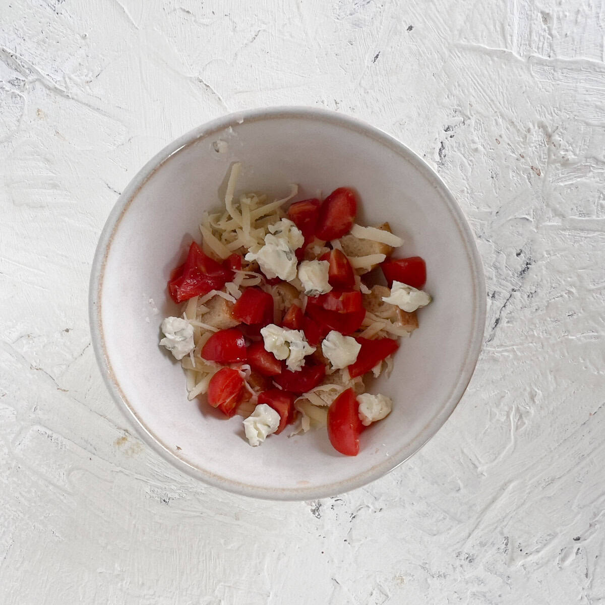A white bowl contains shredded cheese, chopped cherry tomatoes, and small dollops of cream cheese on a textured white surface.