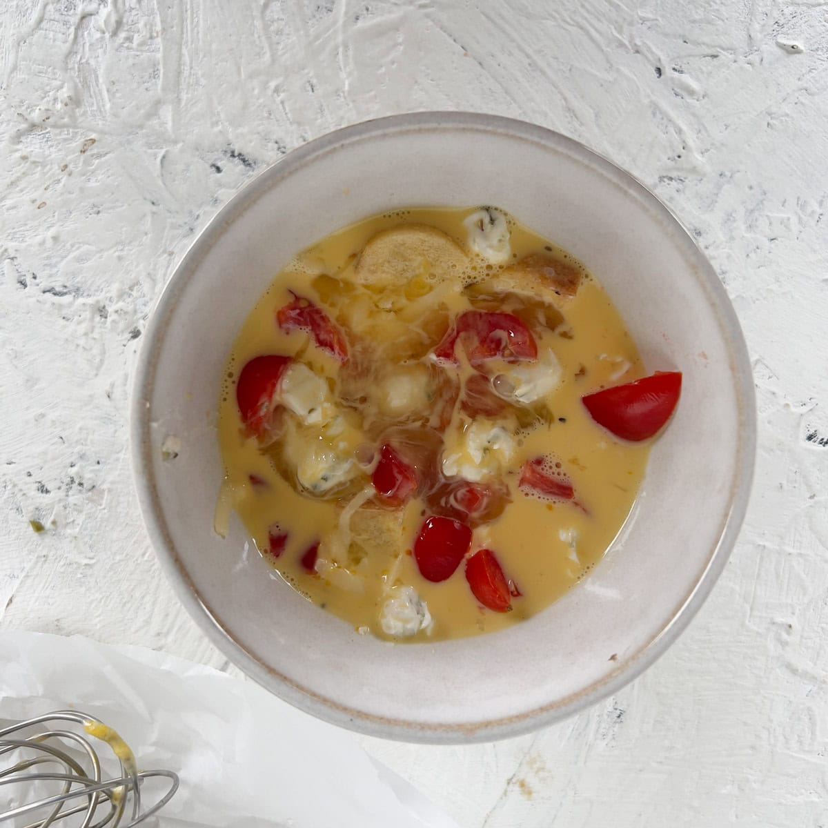 A small metal bowl containing raw beaten eggs, cherry tomato pieces, and chunks of cheese on a white textured surface.