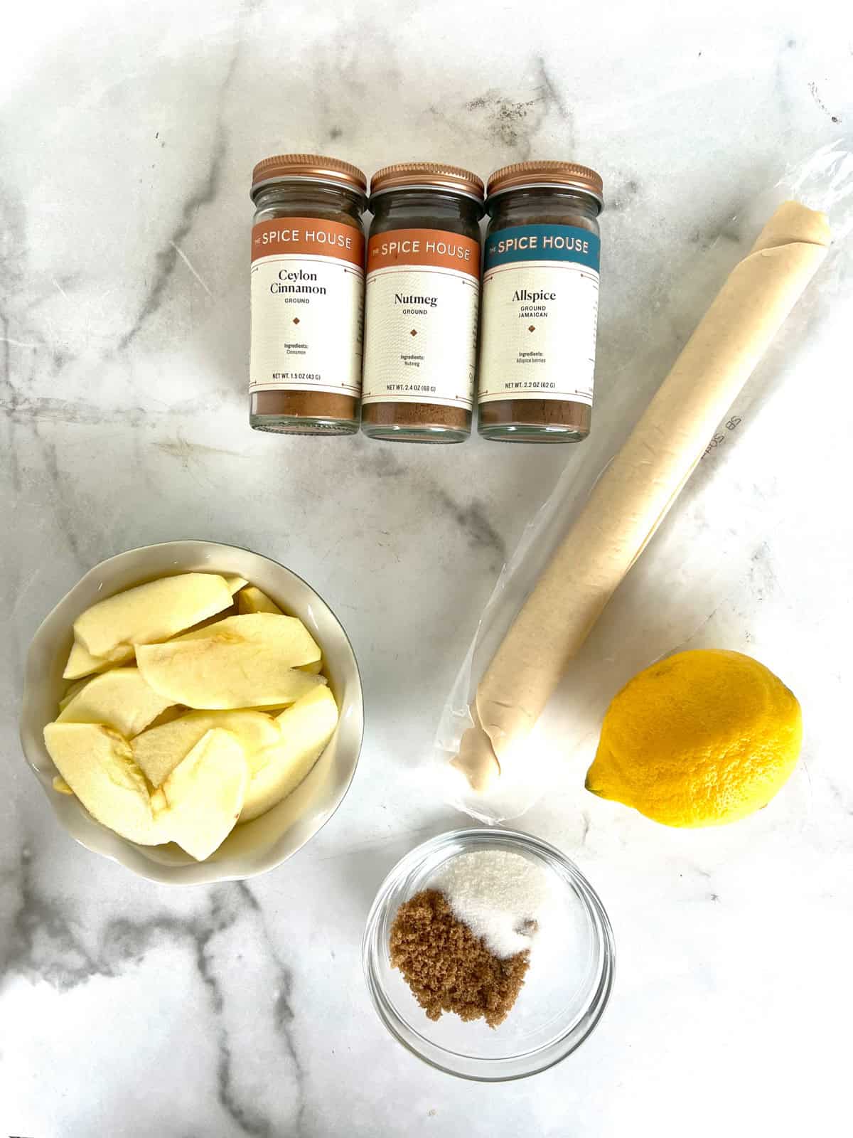 Sliced apples, spices, pie crust, lemon, and sugars arranged on a white marble surface.