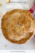 A baked mini apple pie with golden crust and sugar crystals, shown on a white surface with a fork nearby.