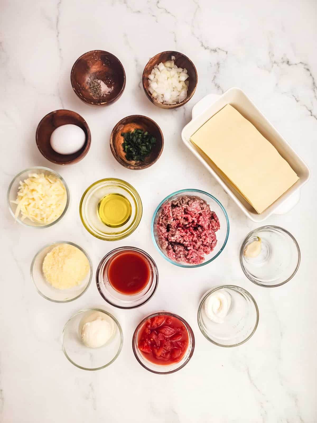 Ingredients for lasagna, including tofu, ground meat, cheese, tomato sauce, egg, onion, and seasonings on a counter.