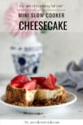 A small cheesecake topped with sliced strawberries sits on a plate, with a vase and napkin in the background.