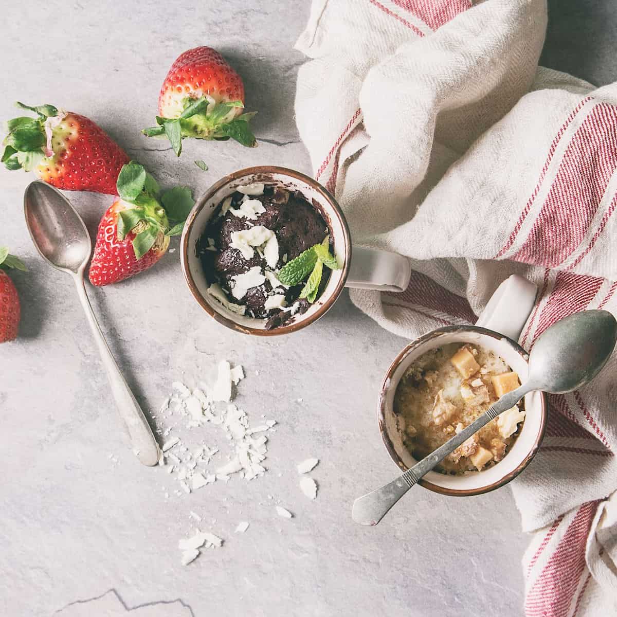 Two mugs with desserts, one chocolate and one vanilla, garnished with shredded coconut and mint, are placed on a light surface with strawberries, spoons, and a striped towel nearby.