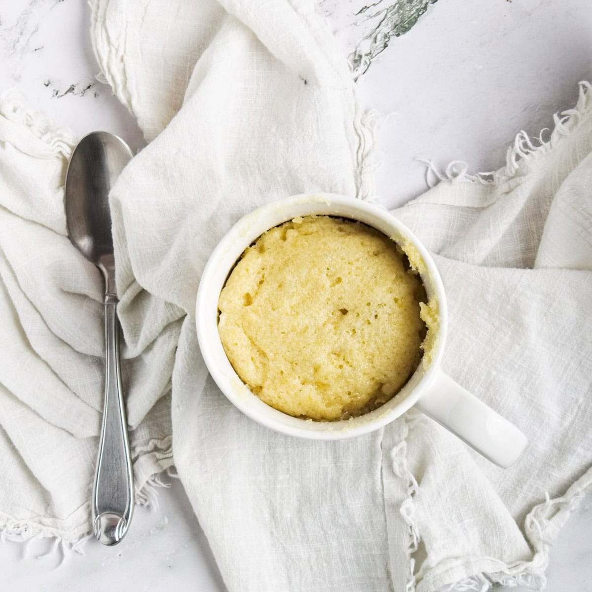 A white mug filled with a baked mug cake sits on a white cloth next to a silver spoon on a marble surface. A white mug filled with a baked mug cake sits on a white cloth next to a silver spoon on a marble surface.