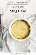 Top-down view of a plain olive oil lemon mug cake in a white mug, placed on a light cloth with a fork beside it. Text on image includes recipe title and website name.