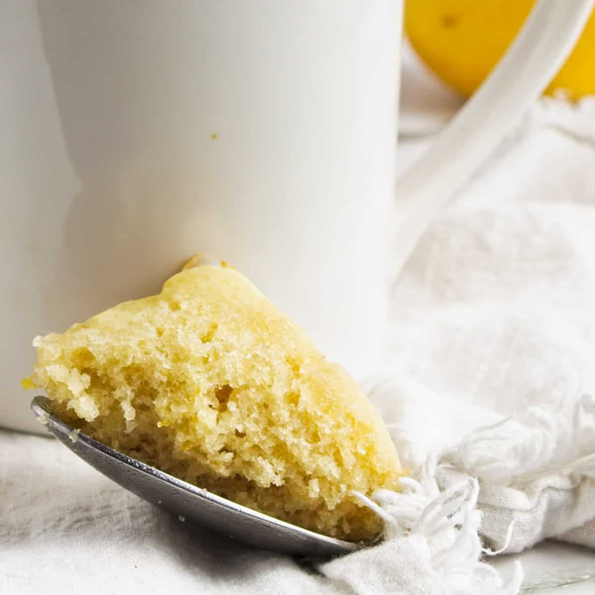 A piece of yellow cake rests on a spoon beside a white mug, with a white cloth and a yellow object visible in the background. A piece of yellow cake rests on a spoon beside a white mug, with a white cloth and a yellow object visible in the background.