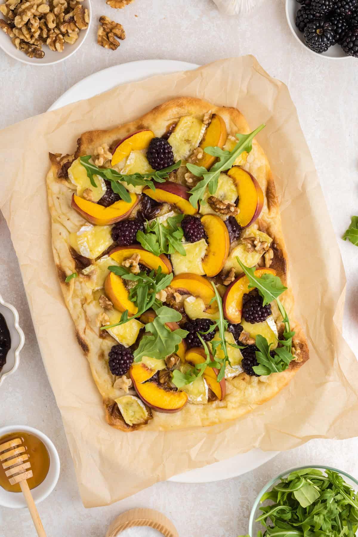 Flatbread topped with peach slices, blackberries, arugula, walnuts, and cheese on parchment paper.