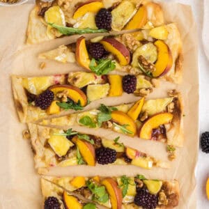 Flatbread topped with cheese, peach slices, blackberries, walnuts, and arugula, cut into wedges on parchment.