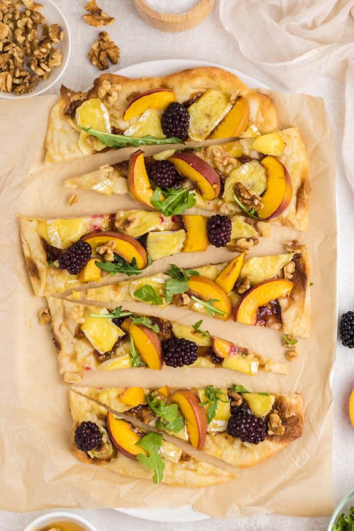 Flatbread topped with cheese, peach slices, blackberries, walnuts, and arugula, cut into wedges on parchment.