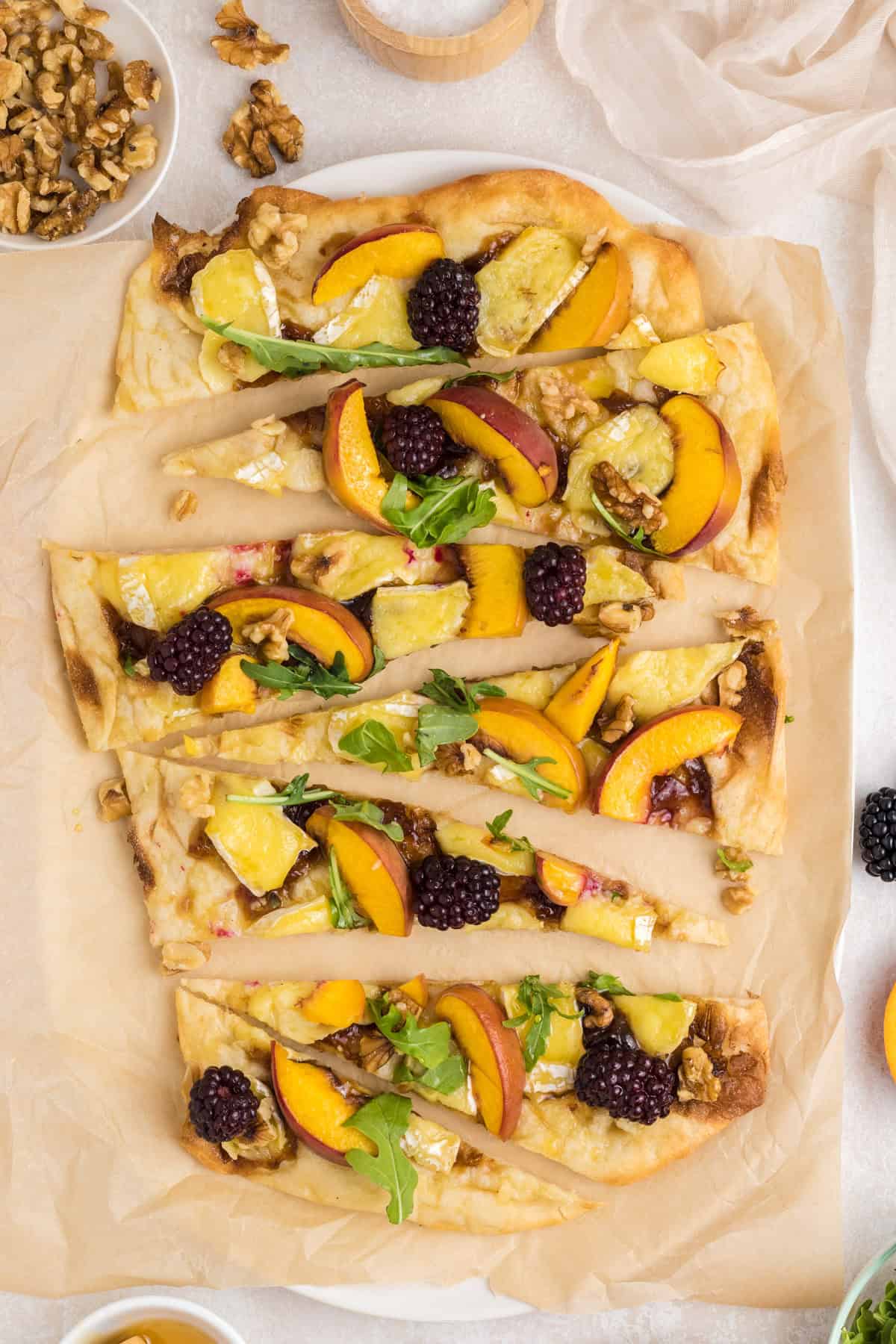 Flatbread topped with cheese, peach slices, blackberries, walnuts, and arugula, cut into wedges on parchment.