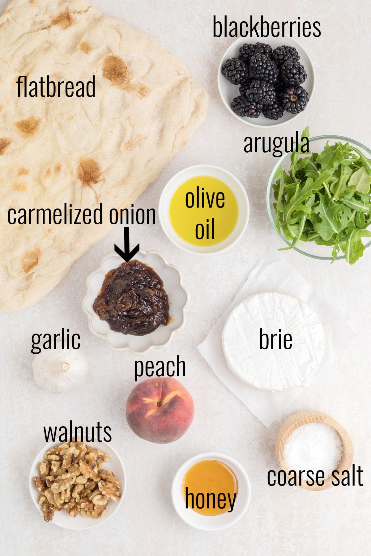 The ingredients for a flatbread pizza, blackberries, brie, arugula, and seasoning are laid out on a table.