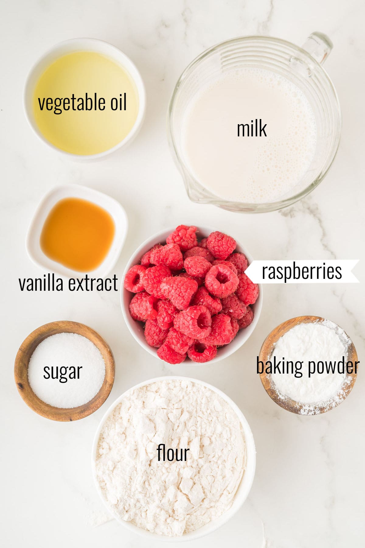 Bowls of milk, vegetable oil, vanilla, raspberries, sugar, flour, and baking powder arranged on a white surface.