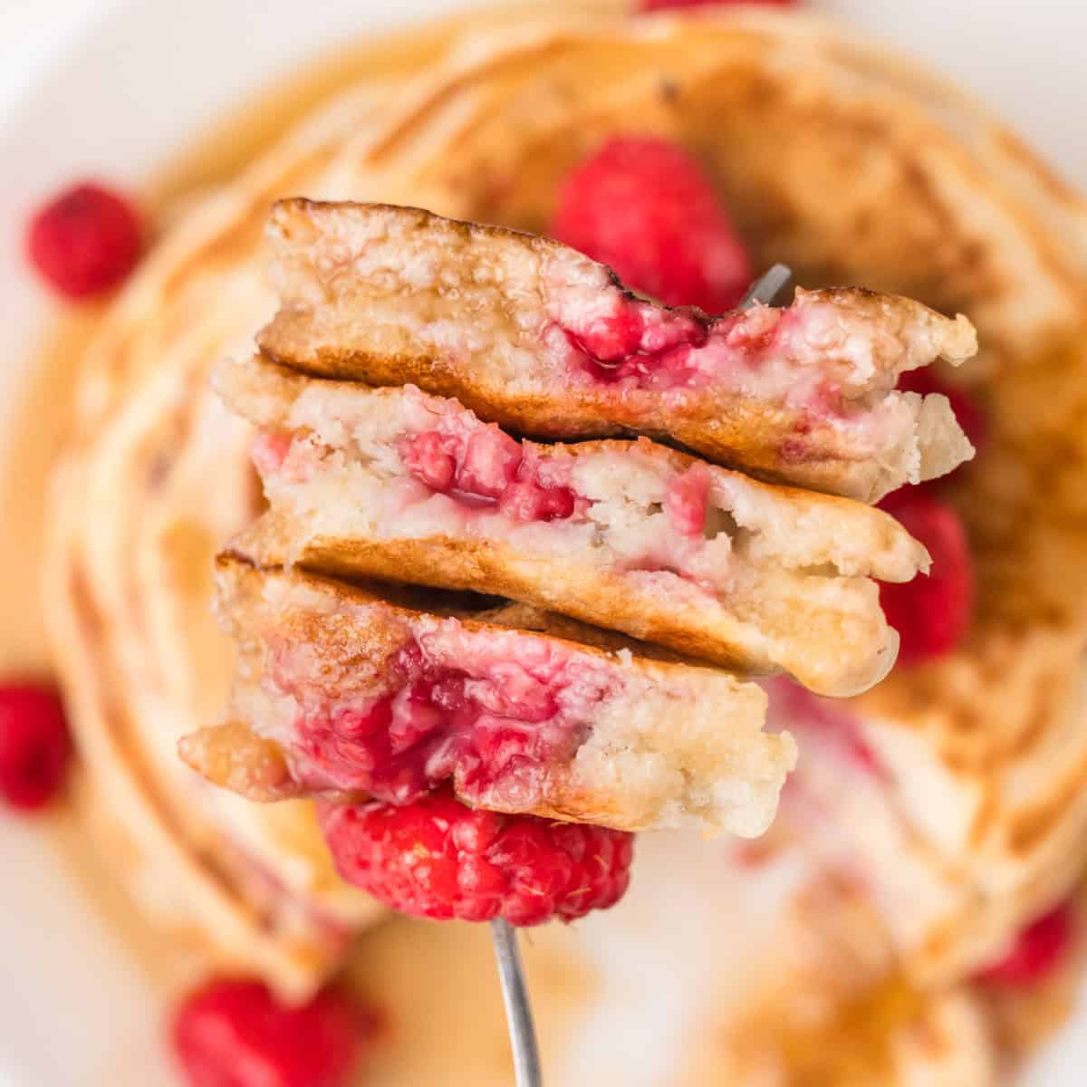 A fork holds a stack of fluffy raspberry pancakes, above a plate with more pancakes and syrup.