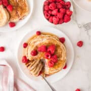 Stack of raspberry pancakes topped with fresh raspberries and syrup, with a fork cutting a piece; fresh raspberries beside.