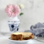 A cheesecake with a slice missing sits on a plate in front of a vase with pink flowers.