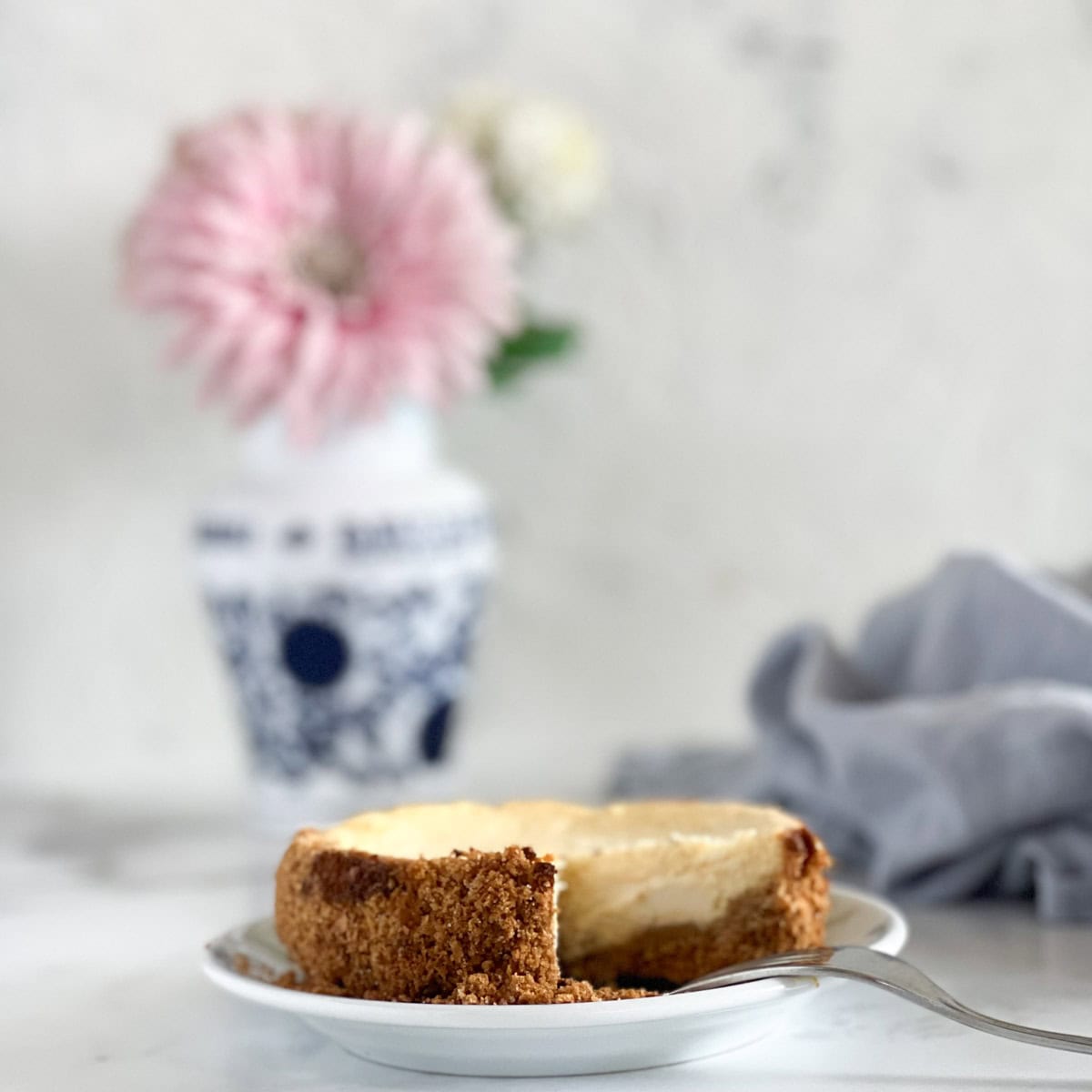 A cheesecake with a slice missing sits on a plate in front of a vase with pink flowers.