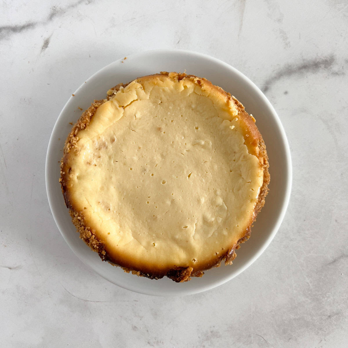 A classic baked cheesecake with a golden crust, served on a white plate against a marble background.