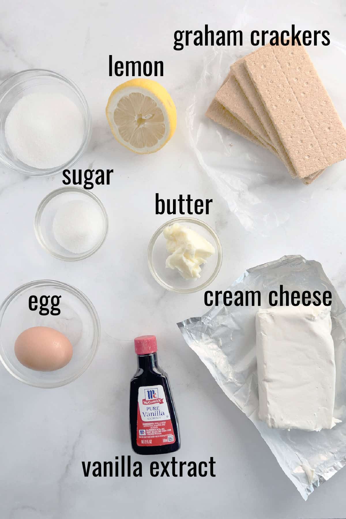 Top view of cheesecake ingredients: graham crackers, lemon, sugar, butter, cream cheese, egg, and vanilla extract.