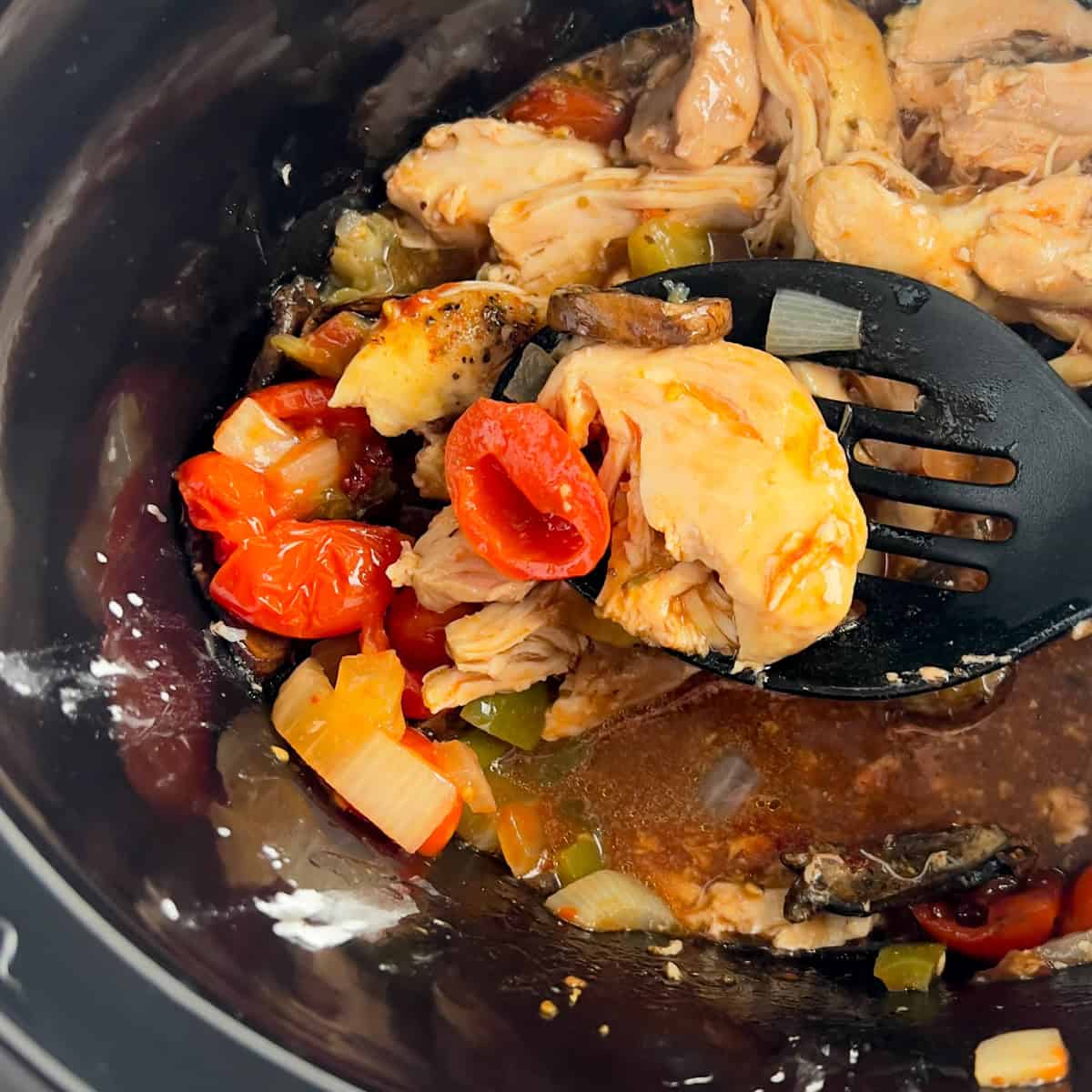 Chicken, tomatoes, peppers, and onions in a slow cooker, with a slotted spoon lifting some pieces.