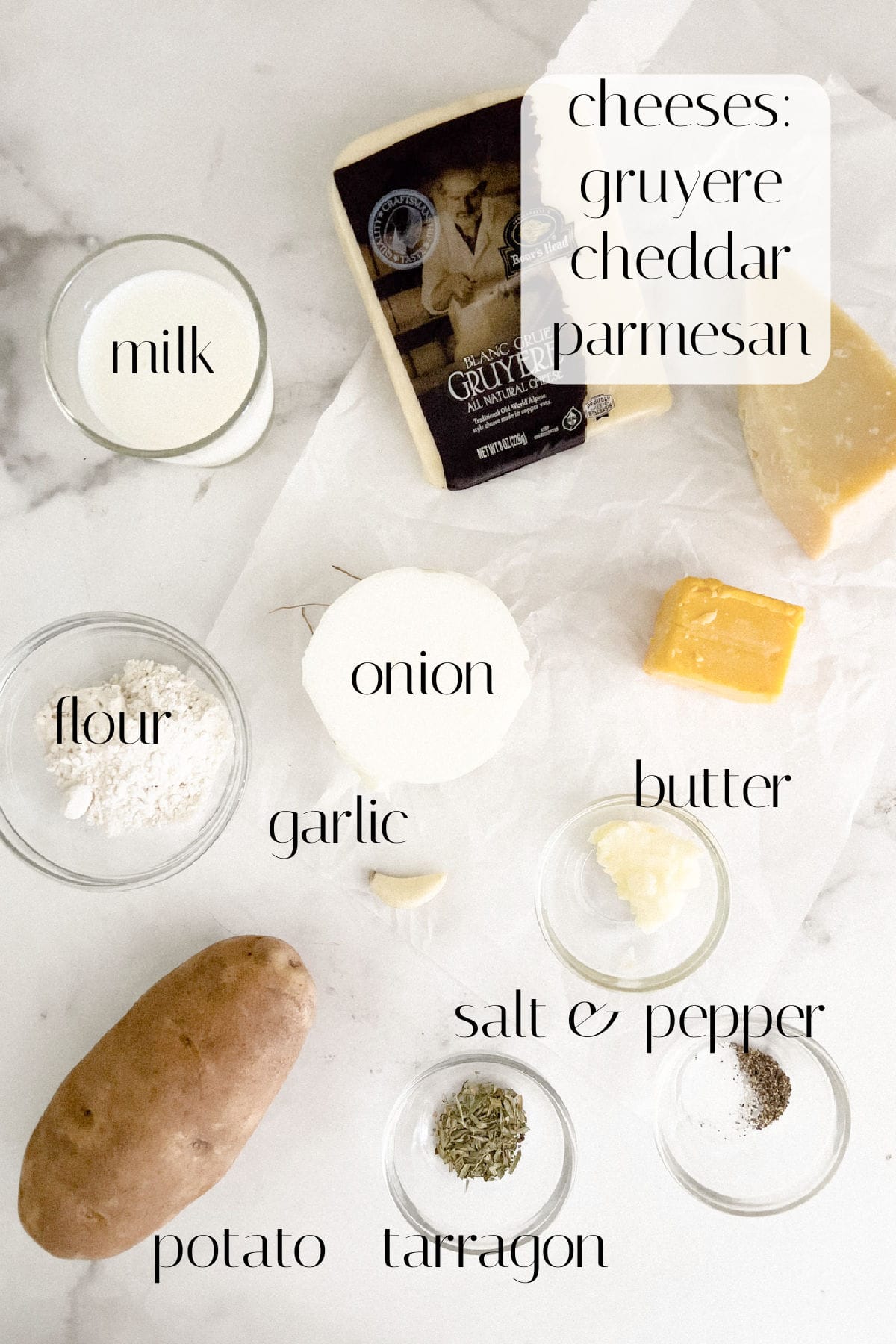 Various ingredients are arranged on a white surface, including milk, cheddar, gruyere, parmesan, flour, onion, butter, garlic, salt, pepper, potato, and tarragon.