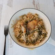 A white skillet contains cooked rice, peas, and seasoned chicken thigh. A black fork is placed to the left of the skillet on a light wood surface.