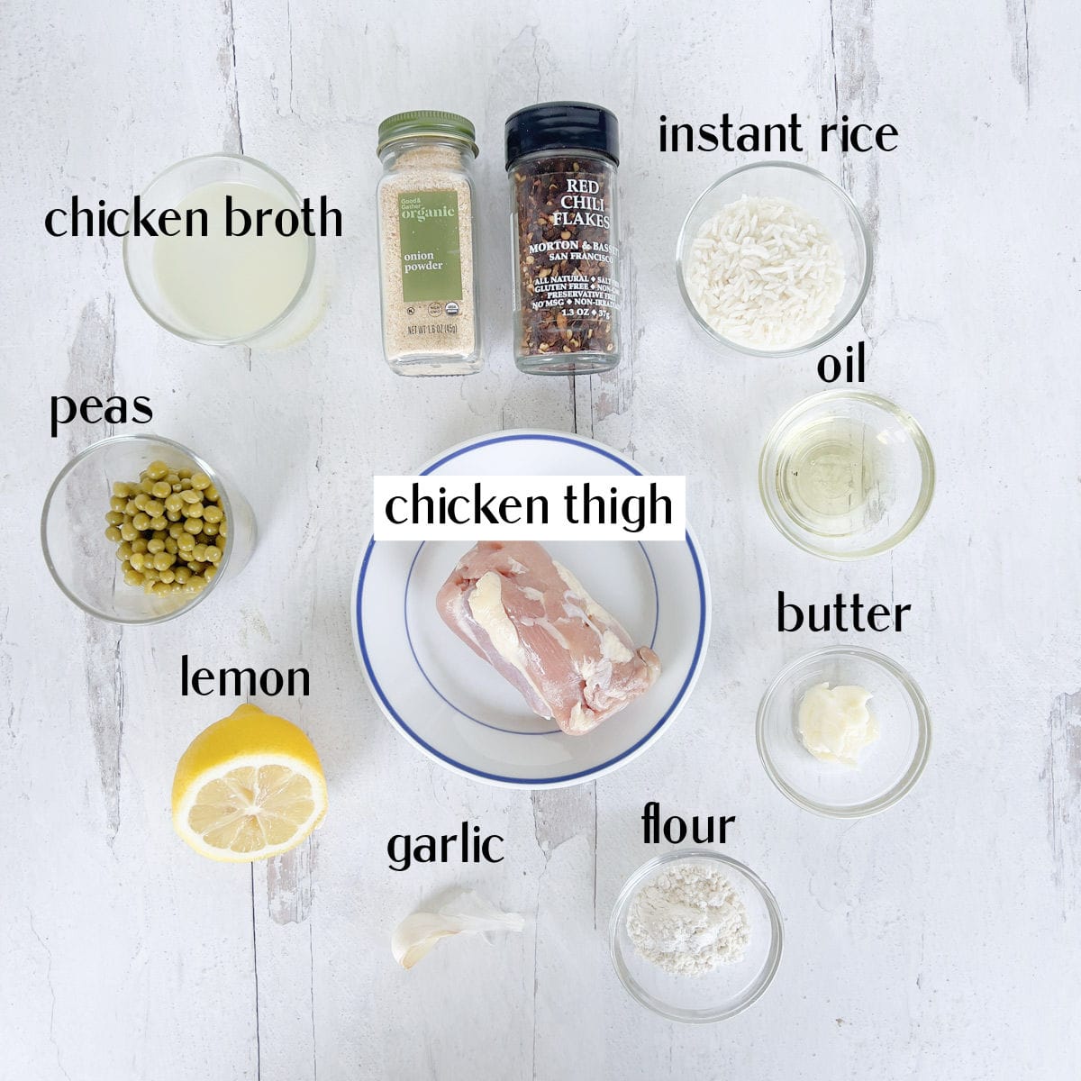 Ingredients for a chicken dish arranged on a white surface, including chicken thigh, peas, lemon, garlic, broth, seasonings, rice, oil, butter, and flour in small bowls.