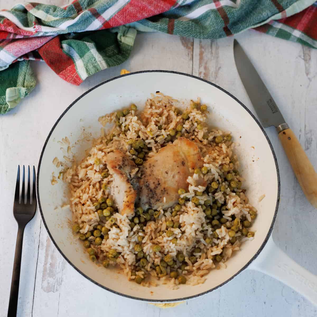 A white skillet contains cooked rice, green peas, and two pieces of seasoned chicken. A fork and knife are placed beside the skillet on a light-colored surface.