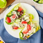 Three lettuce leaves filled with chicken salad, grape tomatoes, sliced cucumbers, and herbs are arranged on a white plate.