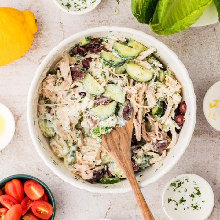 A bowl of creamy chicken salad with cucumbers and olives, surrounded by a lemon, fresh lettuce, grape tomatoes, and seasoning in small bowls.