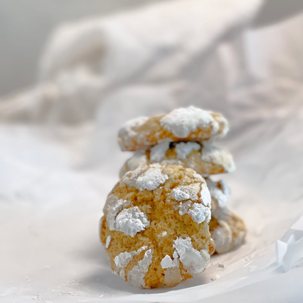 A stack of crinkled cookies dusted with powdered sugar sits on a white, crumpled surface with a soft background.
