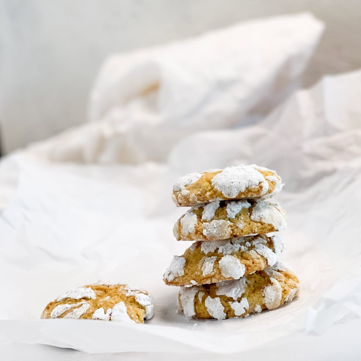 A stack of four lemon crinkle cookies dusted with powdered sugar sits on crumpled white parchment paper, with one cookie lying next to the stack.
