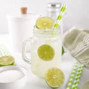 A glass mason jar filled with limeade, ice, and lime slices, with a green striped paper straw, sits on a white surface beside sugar, limes, and kitchen containers.