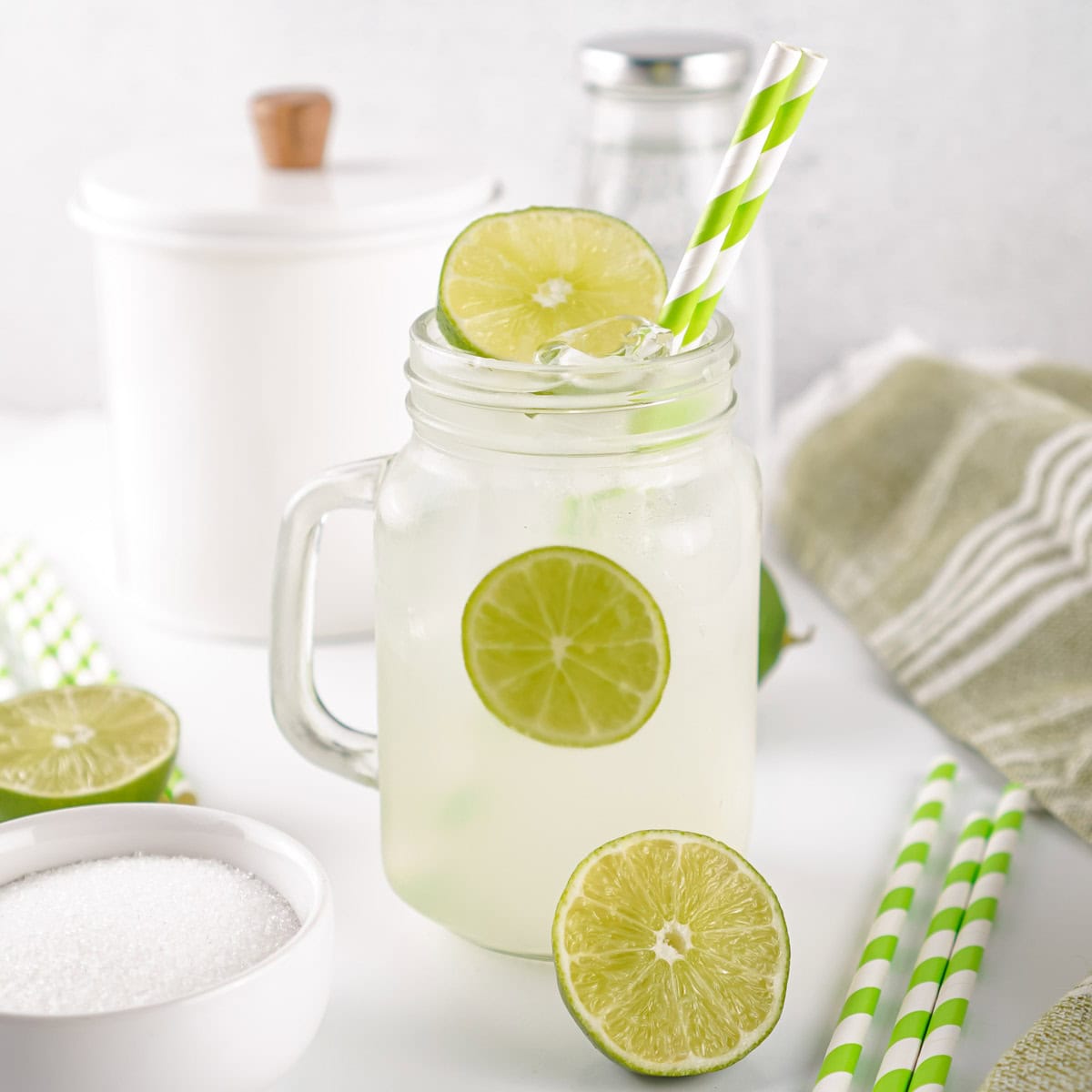 A glass mason jar filled with limeade, ice, and lime slices, with a green striped paper straw, sits on a white surface beside sugar, limes, and kitchen containers.