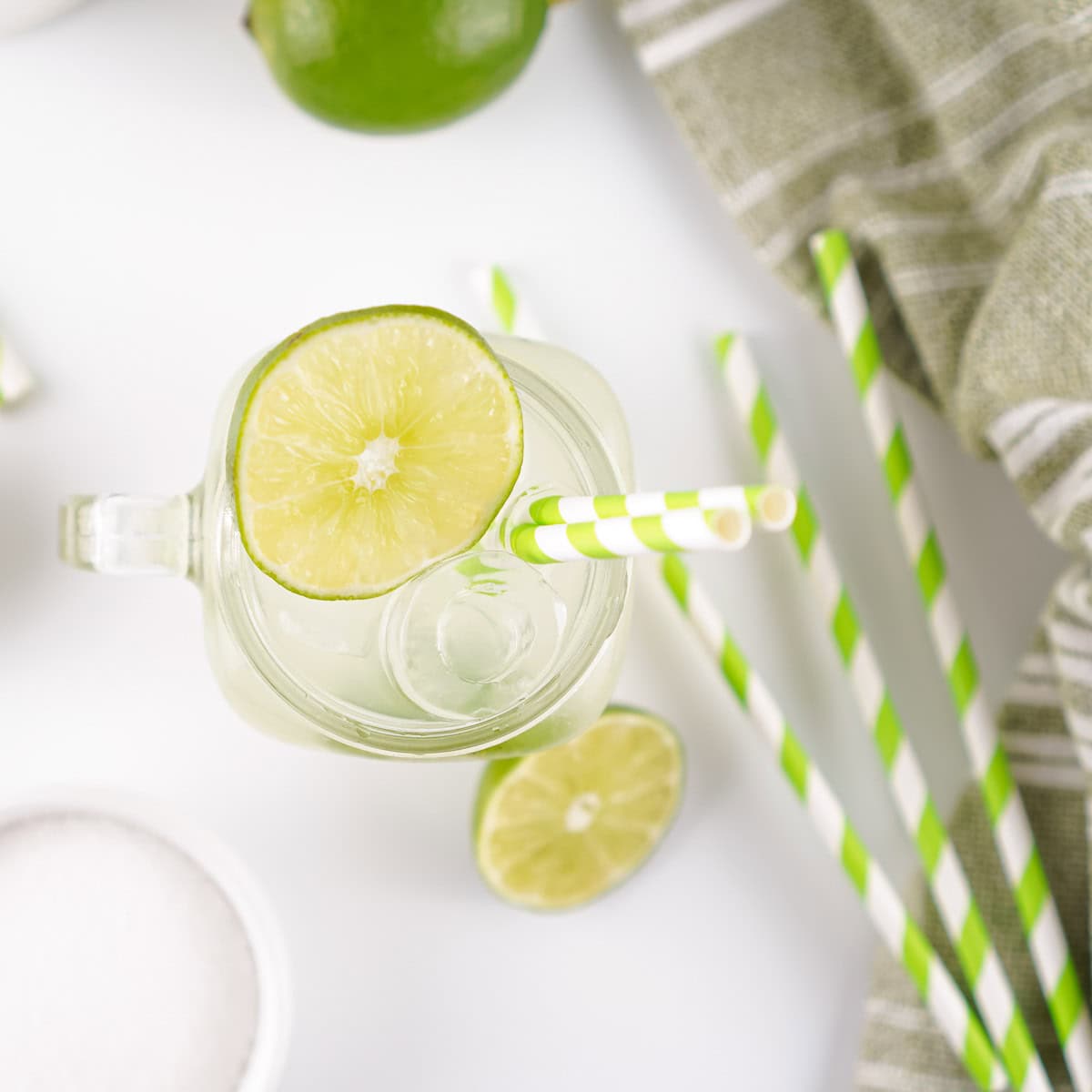 A glass of limeade with ice and a lime slice, green and white striped straws, a halved lime, and a gray cloth nearby on a white surface.