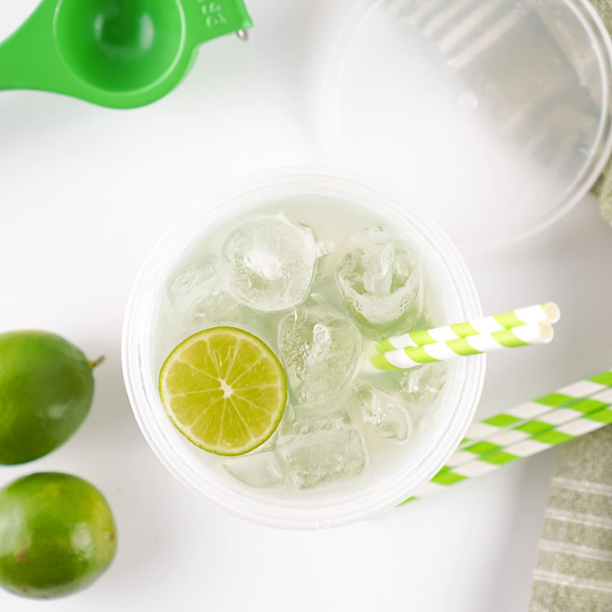 Glass of limeade with ice, a lime slice, and two striped straws in a clear cup, next to whole limes and a green citrus squeezer on a white surface.