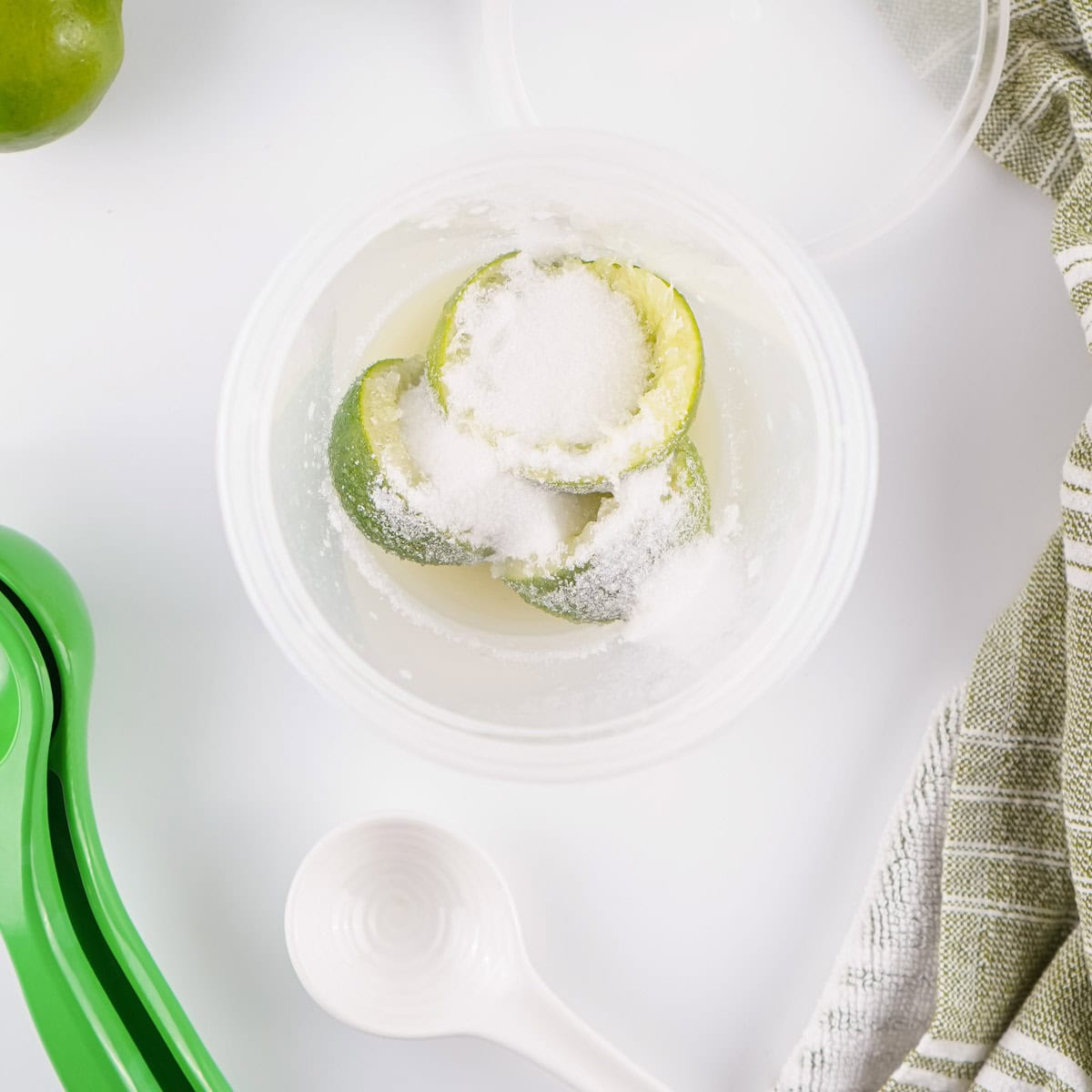 A plastic container with halved limes covered in sugar, a white measuring spoon, a green citrus squeezer, and a striped cloth on a white surface.