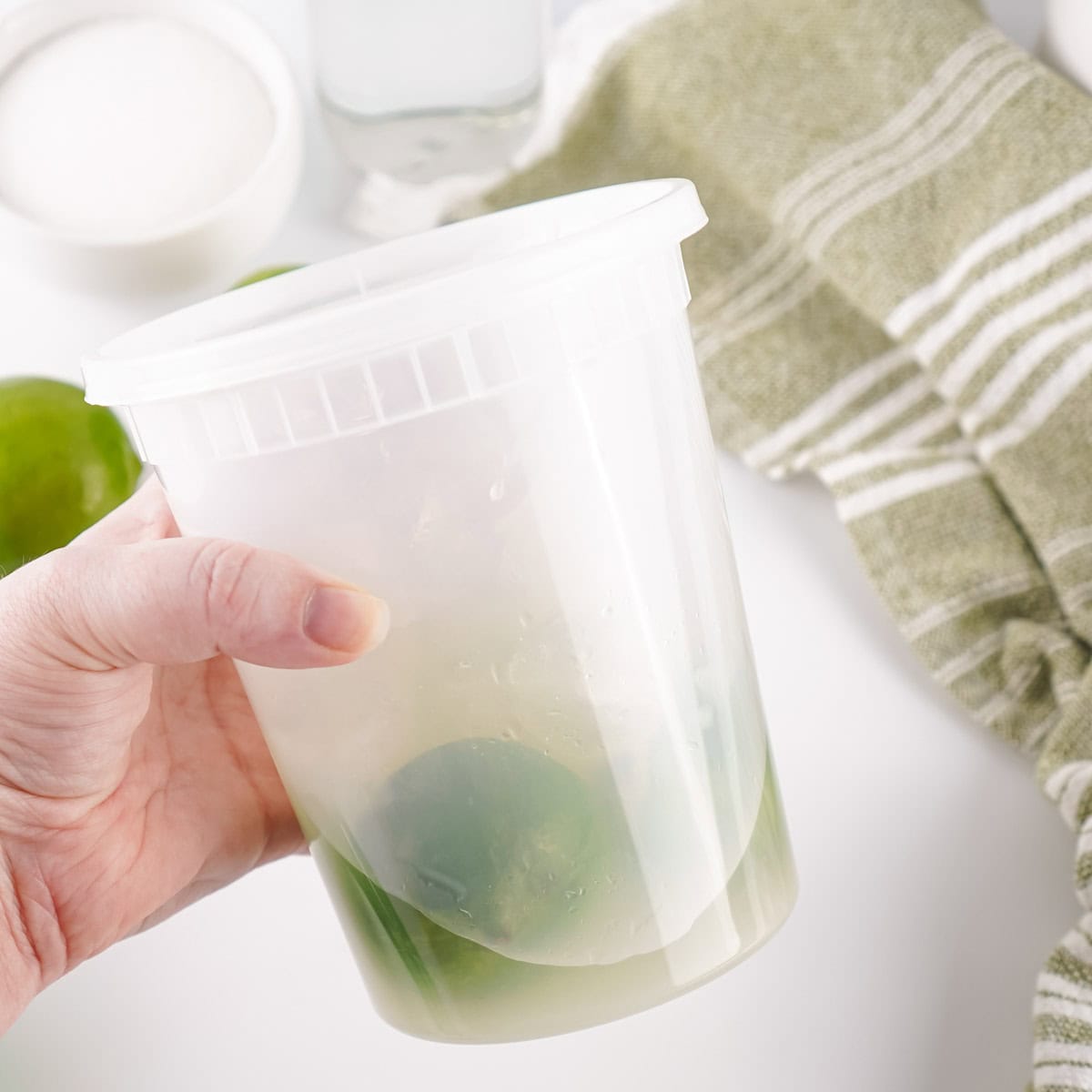 A hand holds a plastic container with whole limes and juice inside; a green striped towel and bowl of sugar are in the background.