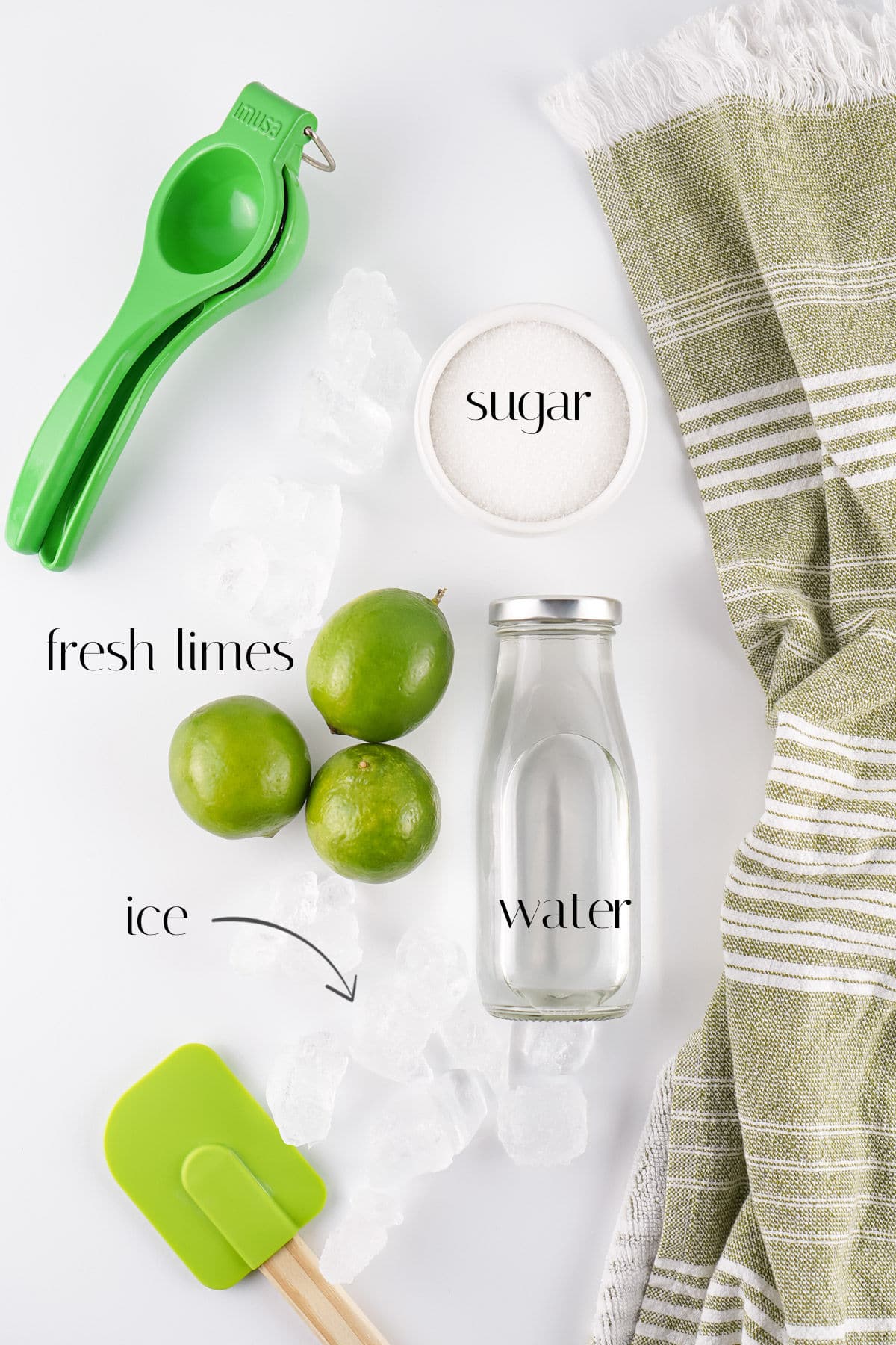 Overhead view of a lime squeezer, three limes, a jar of water, a bowl of sugar, ice cubes, a green spatula, and a green-striped towel on a white surface.