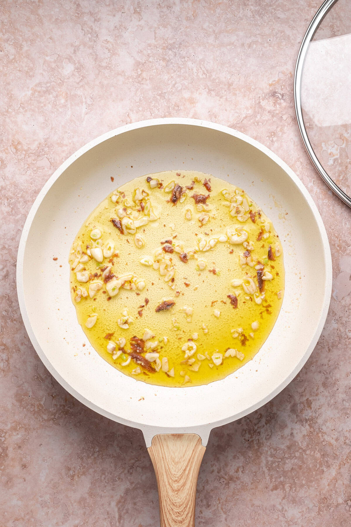 Overhead view of a frying pan with chopped garlic and bits of bacon sautéing in oil on a pink countertop; a glass lid is partially visible.
