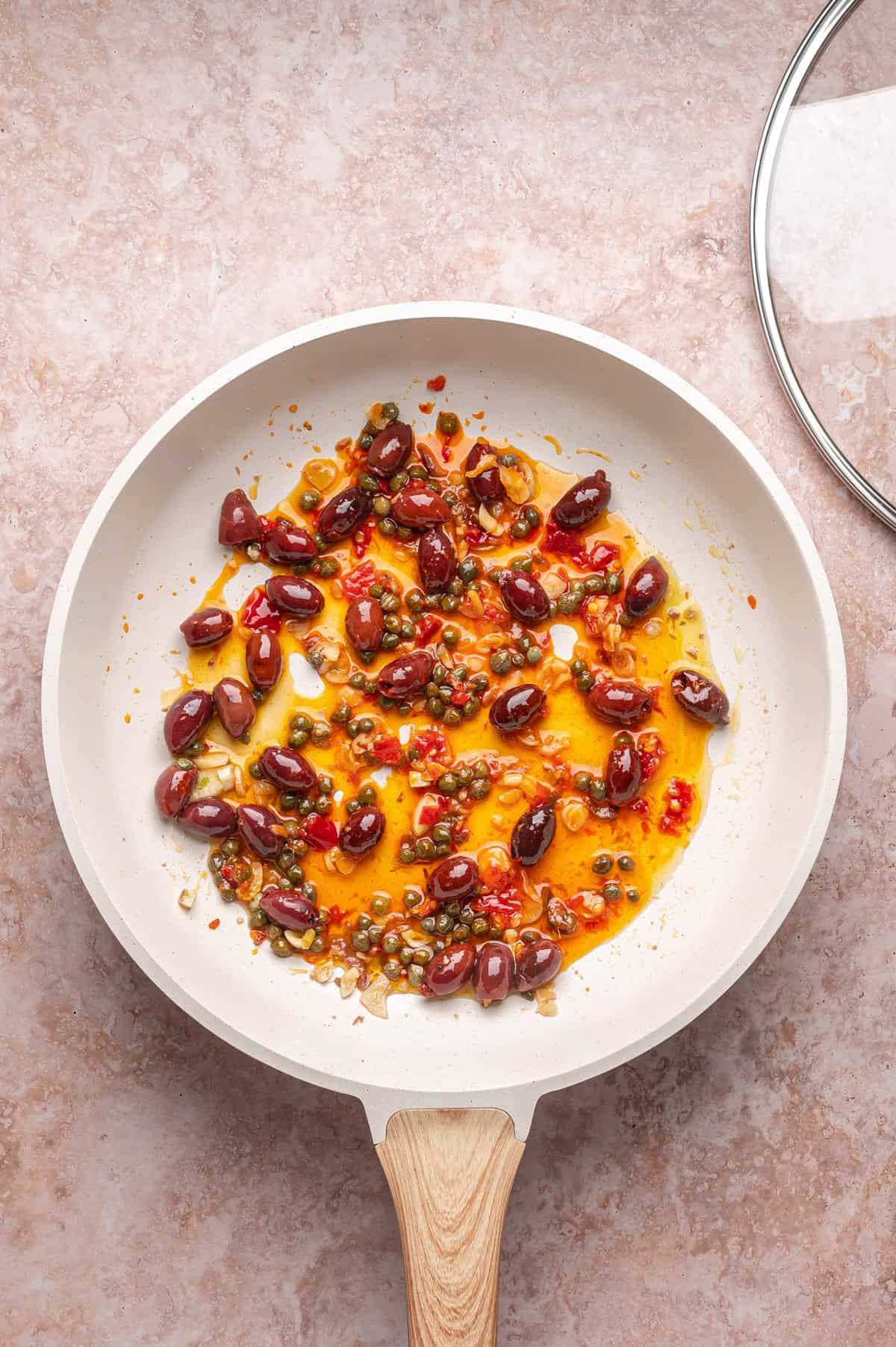 A white frying pan with sautéed Kalamata olives, capers, minced garlic, and red pepper flakes in oil on a pink marble surface. The glass lid is partially visible to the side.