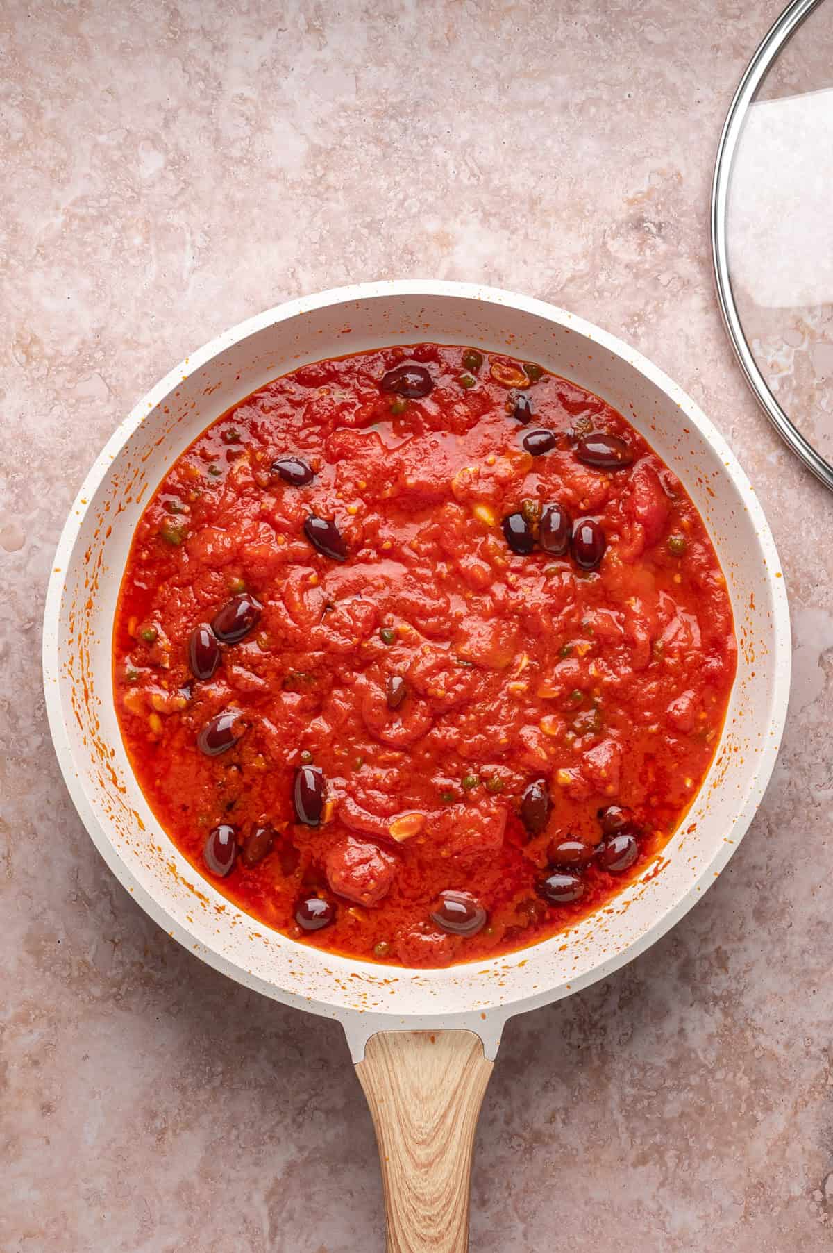 A white frying pan filled with tomato sauce and black olives sits on a beige countertop next to a glass lid.