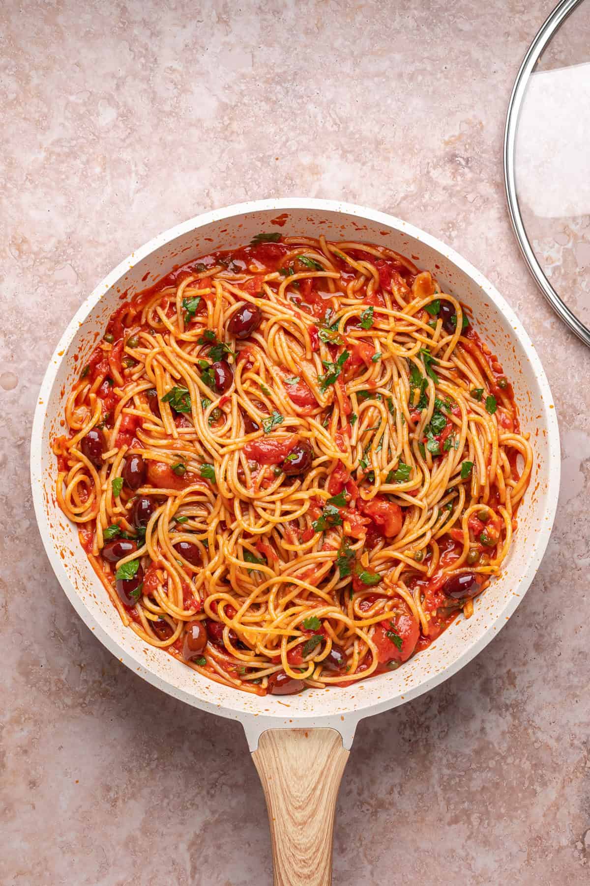 A pan of spaghetti in tomato sauce with olives and fresh herbs sits on a light-colored countertop next to a glass lid.