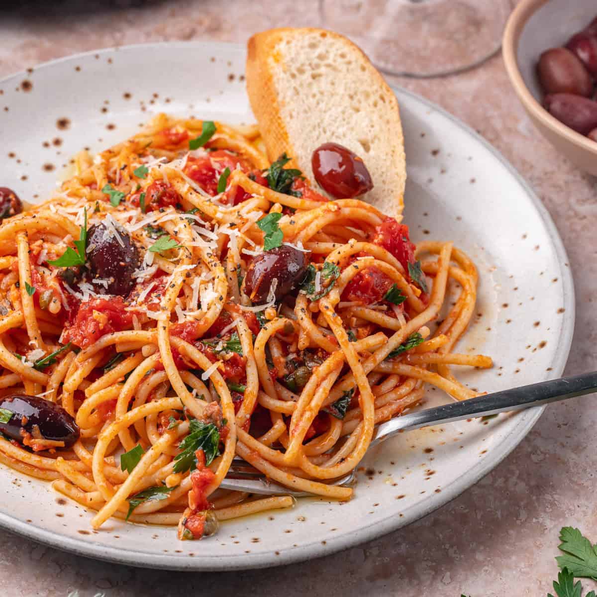 A plate of spaghetti with tomato sauce, black olives, grated cheese, and parsley, served with a slice of bread. A fork rests on the plate.