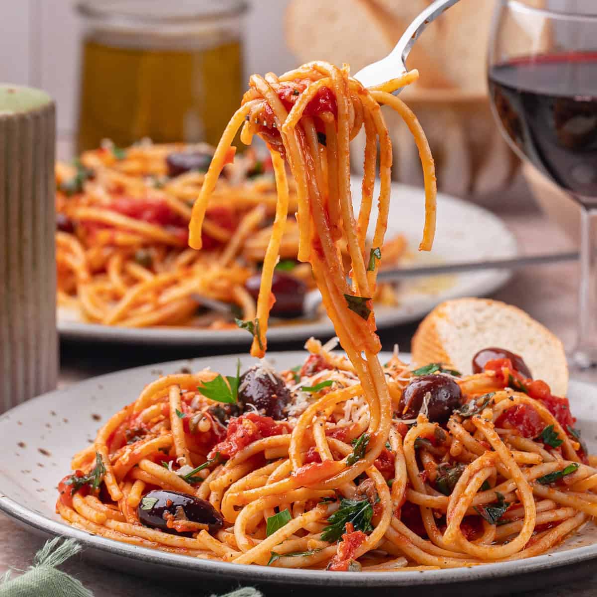 A plate of spaghetti with a puttanesca sauce, black olives, and herbs, served with a slice of bread; a fork lifts a portion. A glass of red wine and a jar are in the background.