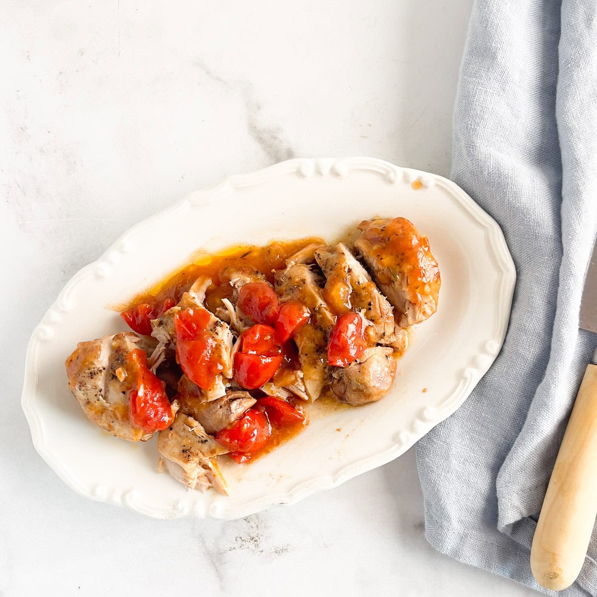 Sliced cooked chicken with tomato sauce on a white plate, next to a light blue napkin and wooden-handled knife.